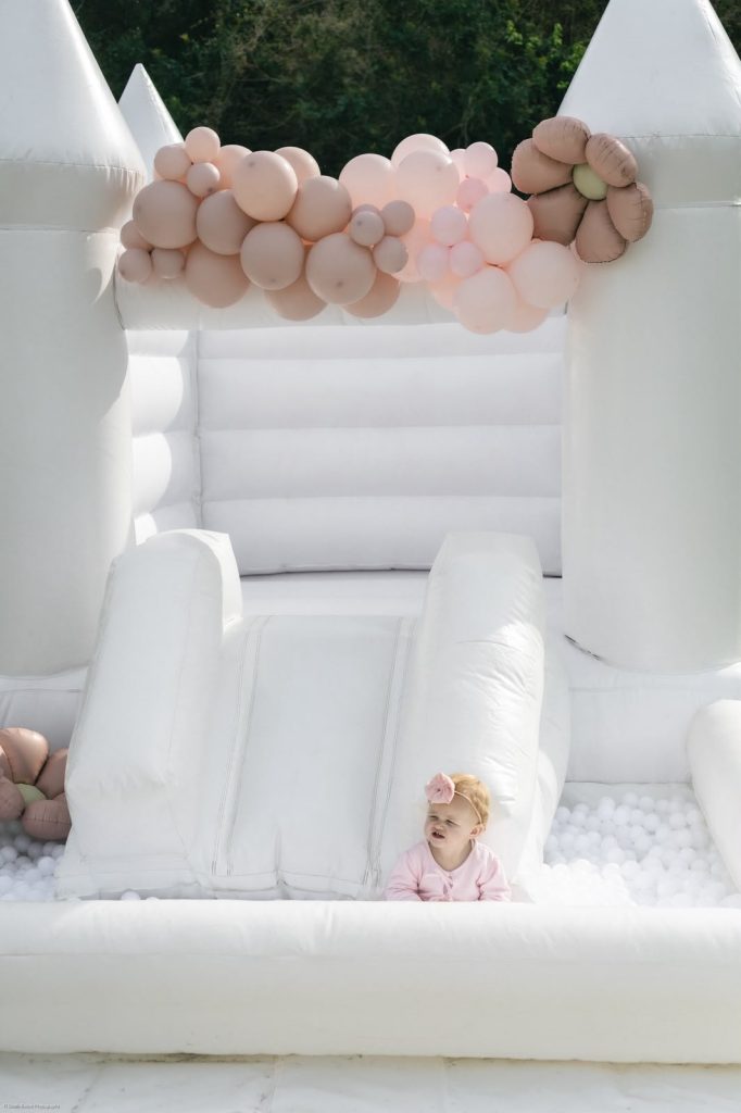 White jumping castle with a blush balloon garland, slide and white ball pit