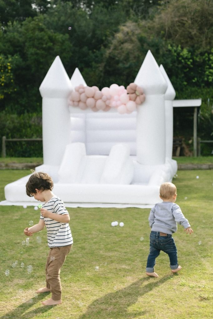 Two children chasing bubbles on the lawn in front of the white jumping castle